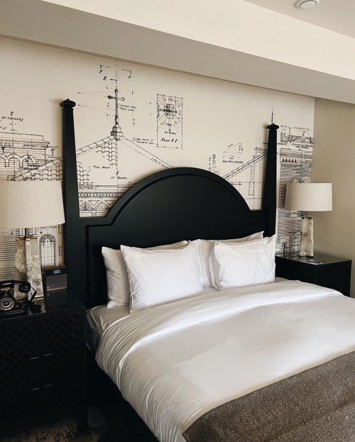 A stylish hotel room featuring a bed with white linens and architectural sketches on the wall behind it.