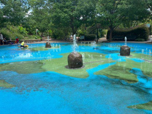 A colorful splash pad with water features and rocks, surrounded by greenery and children playing.