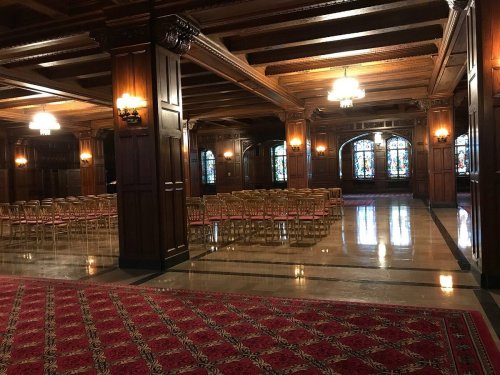 Elegant interior of a hall with wooden beams, stained glass windows, and rows of empty chairs on a patterned carpet.