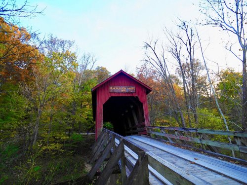 A red covered bridge surrounded by autumn trees and a wooden walkway leading to it.