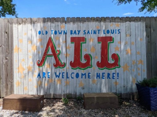 Colorful mural on a wooden fence reads "ALL ARE WELCOME HERE!" in Old Town Bay Saint Louis.