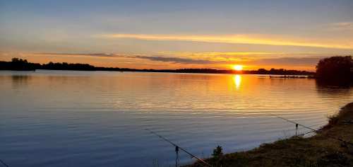 A serene sunset over a calm lake, with fishing rods positioned along the shore.