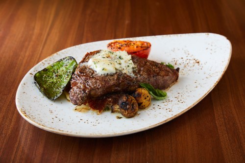 A perfectly cooked steak topped with herb butter, served with grilled vegetables and a tomato on a white plate.