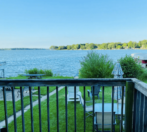 A serene lakeside view with green grass, lounge chairs, and trees under a clear blue sky.