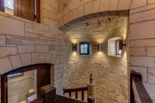 A spiral staircase with stone walls and warm lighting, featuring small windows and wooden railings.