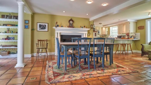 A cozy dining area with a wooden table, blue chairs, and a fireplace, featuring warm colors and tiled flooring.