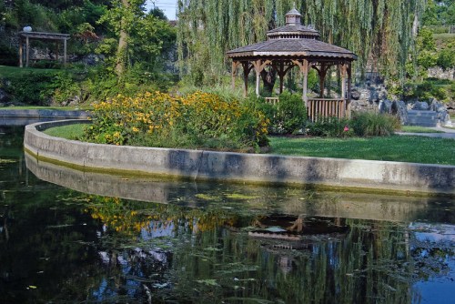 A serene park scene featuring a gazebo surrounded by flowers, reflecting in a calm pond with lush greenery.