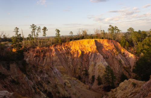 A colorful canyon landscape with steep, layered cliffs and trees against a clear sky.