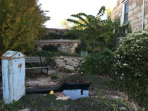 A serene garden scene featuring a pond, stone steps, a bench, and lush greenery surrounding a rustic blue structure.