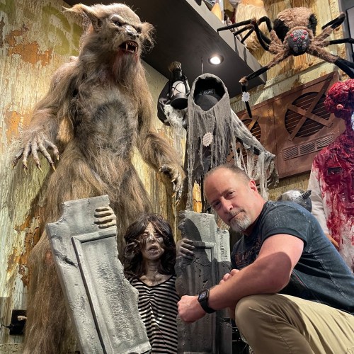 A man poses with creepy horror-themed decorations, including a werewolf and a ghostly figure, in a dimly lit room.