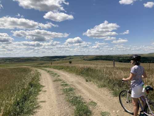 A cyclist stands on a dirt path, overlooking rolling hills and a blue sky filled with fluffy clouds.