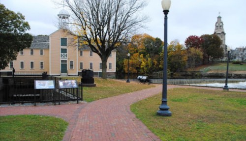 A brick pathway leads past a historic building and trees with autumn leaves, near a river and a church steeple in the background.