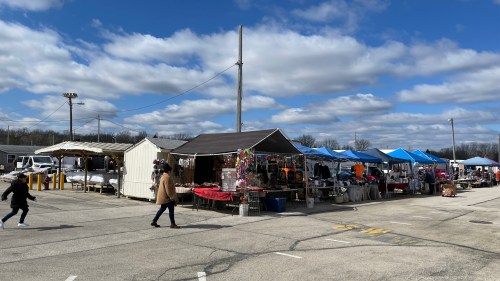 A bustling outdoor market with colorful tents and vendors under a partly cloudy sky. People are walking around.