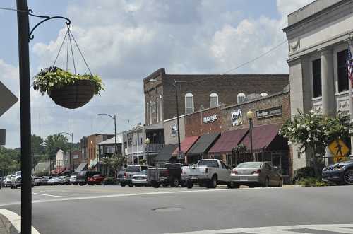 A street view of a small town with shops, parked cars, and hanging flower baskets under a partly cloudy sky.