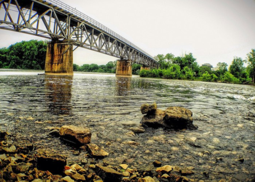 A steel bridge spans a river, with rocky shorelines and lush greenery in the background under a cloudy sky.