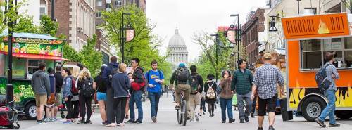 A busy street scene with food trucks and people walking, featuring a view of a dome building in the background.