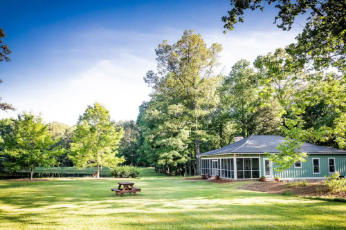 A serene landscape featuring a green lawn, a picnic table, and a house surrounded by trees under a clear blue sky.