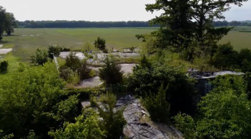 Aerial view of overgrown ruins surrounded by lush greenery and open fields in the background.