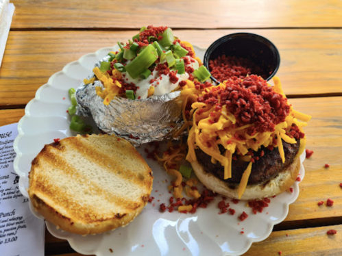 A plate with a loaded baked potato topped with cheese and green onions, and a burger with cheese and bacon bits, on a wooden table.