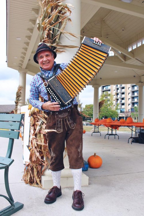 A man in traditional attire holds an accordion, smiling at an outdoor event with pumpkins in the background.