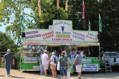 A colorful food stand offers frozen daiquiris, lemonade, corn dogs, and funnel cakes, with customers lined up to order.