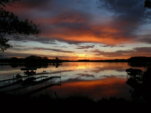 A serene lake at sunset, with vibrant orange and purple skies reflecting on the water and silhouettes of boats.