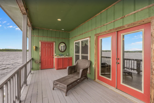 A cozy porch with green walls, red doors, and a view of the water, featuring a lounge chair and a small kitchenette.