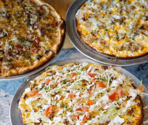 Three pizzas on metal trays: one with meat and herbs, one with cheese and toppings, and one with lettuce and tomatoes.