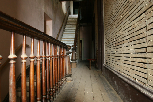 A dimly lit hallway with a wooden railing, leading to a staircase and a textured wall.