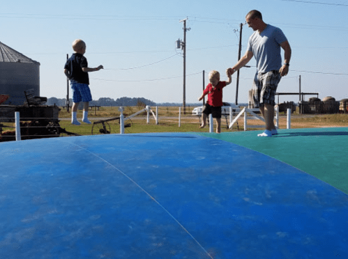 A man holds hands with a child while another child jumps on a colorful inflatable surface in a rural setting.