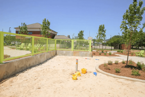 A sunny playground with a sandy area, green fencing, and scattered toys, surrounded by trees and seating.