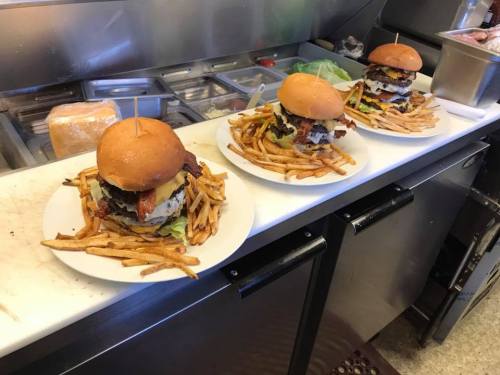 Three large burgers stacked high with toppings, served on plates with a side of crispy French fries.