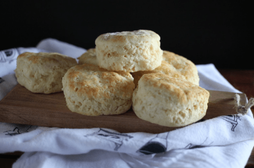 A stack of golden, flaky biscuits on a wooden board, resting on a white cloth.