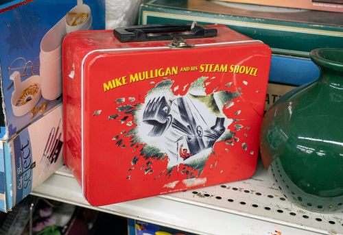 A vintage red lunchbox featuring artwork from "Mike Mulligan and His Steam Shovel," displayed on a shelf.