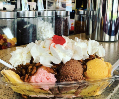 A colorful ice cream sundae with three scoops, whipped cream, chocolate syrup, and a cherry on top, served in a bowl.