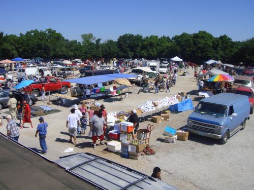A bustling outdoor market with vendors, cars, and shoppers under a clear blue sky. Tents and tables are set up throughout.