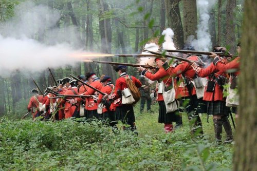 A group of soldiers in red uniforms firing muskets in a forest during a reenactment. Smoke billows around them.
