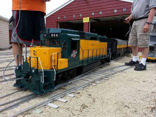 A model train engine in green and yellow, parked on tracks with two people nearby, in front of a red garage.
