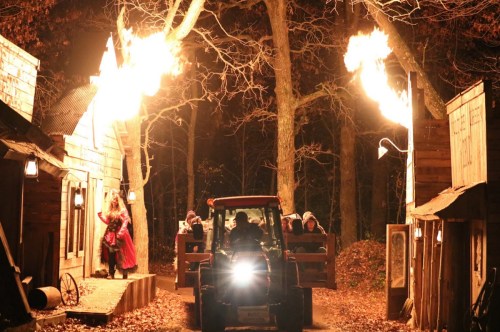 A tractor drives through a dark, wooded area with flames shooting up from either side, illuminating a rustic village.