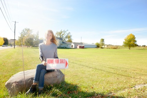 A woman sits on a rock holding a "Home for Sale" sign in a grassy area with trees and a house in the background.