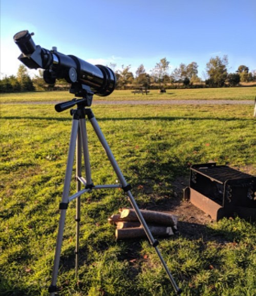A telescope set up on a grassy field, with a fire pit nearby and trees in the background under a clear blue sky.