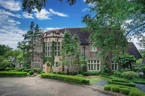 A large, elegant brick house surrounded by lush greenery and a well-maintained garden under a blue sky.