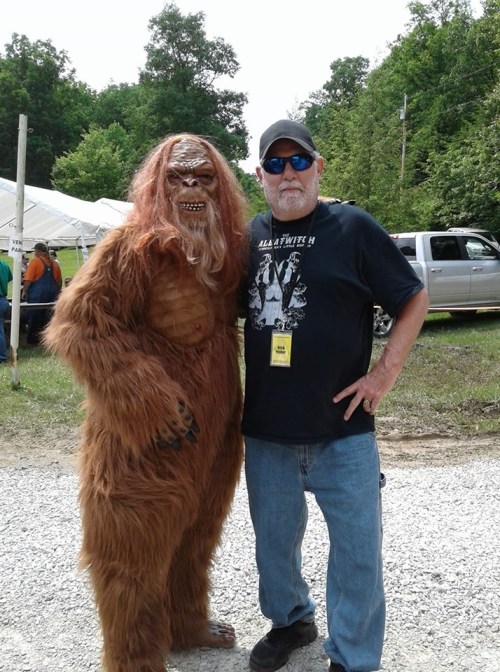A person in a Bigfoot costume poses with a man wearing sunglasses and a black t-shirt at an outdoor event.