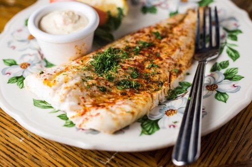 A seasoned fish fillet garnished with herbs, served with a side of sauce and a lemon wedge on a floral plate.