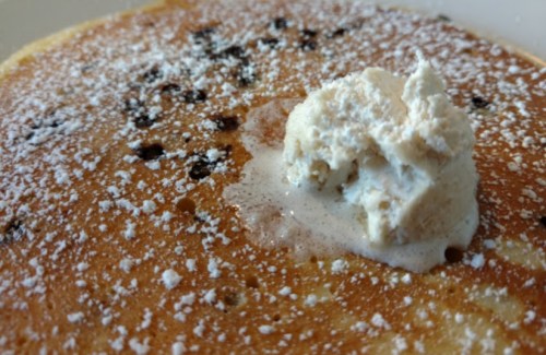 Close-up of a fluffy pancake topped with whipped cream and dusted with powdered sugar, featuring chocolate chips.