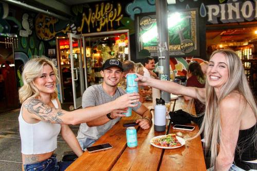 Three friends cheer with drinks at a lively outdoor table, enjoying food and a vibrant atmosphere.