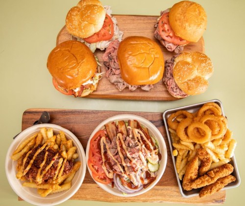 A wooden platter with three burgers, fries, onion rings, and a bowl of loaded fries topped with sauce and meat.