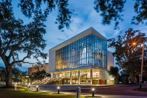 Modern glass building illuminated at dusk, surrounded by trees and outdoor lighting.