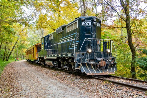 A black locomotive with a yellow train car on a gravel track surrounded by colorful autumn trees.