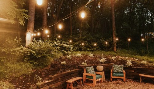 Cozy outdoor seating area with string lights, surrounded by greenery and wooden landscaping.